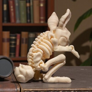Puede incluir: Una escultura de color crema de un esqueleto de conejo en posici&oacute;n agachada. La escultura detallada muestra la caja tor&aacute;cica, la columna vertebral y los huesos de las piernas. Las orejas del conejo est&aacute;n erguidas y la escultura est&aacute; sobre una superficie oscura.