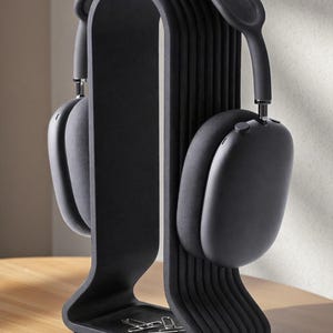 May include: A black headphone stand with a pair of black headphones. The stand has a curved design with a ribbed back and a small tray at the base. The headphones are resting on the stand, showcasing their design.
