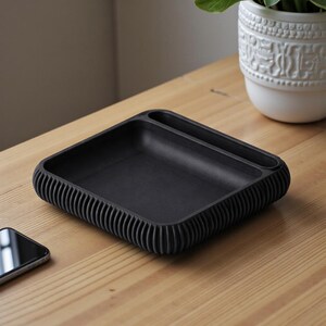 May include: A black desk organizer tray with a ribbed edge design. The tray has a rectangular shape with a small, built-in compartment. A smartphone is visible on the wooden surface next to the tray. The tray is designed to hold small items.