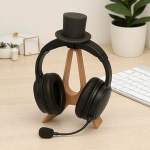 May include: Black over-ear headphones with a microphone, resting on a wooden stand shaped like an A-frame. A small, black top hat sits atop the stand. The headphones are on a light-colored wooden desk, with a plant and keyboard in the background.