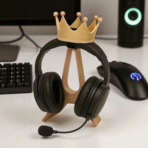 May include: A black headset with a microphone rests on a wooden headphone stand, topped with a gold-colored crown. A black computer mouse and speaker are in the background. The setup is on a white desk.