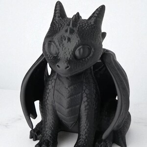 May include: A black dragon figurine with large, expressive eyes, wings, and a textured surface. The dragon is sitting, with its wings spread slightly. The figurine has a detailed, scaled appearance, and is a fantasy-themed decorative item.