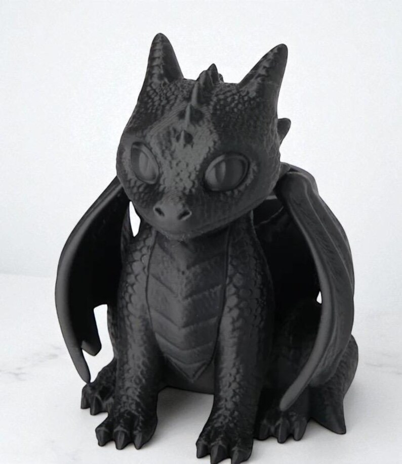 May include: A black dragon figurine with large, expressive eyes, wings, and a textured surface. The dragon is sitting, with its wings spread slightly. The figurine has a detailed, scaled appearance, and is a fantasy-themed decorative item.