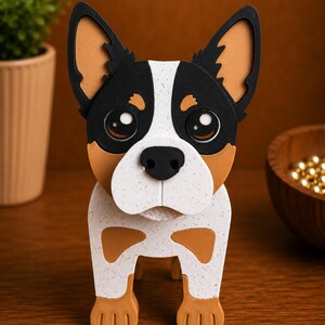 May include: A handcrafted dog figurine with a tri-color design. The dog has a black head with tan ears, a white face and chest, and tan paws. The figurine is made of layered materials and stands on a wooden surface.