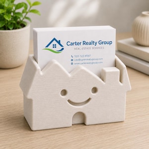 May include: A beige house-shaped business card holder with a smiling face cutout. The holder contains a stack of white business cards with the text "Carter Realty Group" and contact information. The holder sits on a light wood surface.