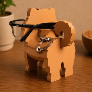 May include: A light brown, dog-shaped eyeglasses and key holder. The holder has a slot for glasses and a ring for keys. Black eyeglasses are resting on the holder. The holder is made of wood and has a textured appearance.