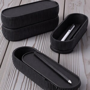 May include: Black, ribbed, oval pencil cases with lids. One case is open, revealing a pencil, and another contains a pen. The cases are stacked and arranged on a light wood-grain surface. The cases have a modern, minimalist design.