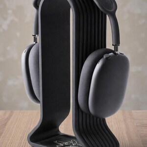 May include: Black over-ear headphones resting on a black headphone stand. The stand has a curved design with a small tray at the base. The headphones and stand are on a wooden surface.