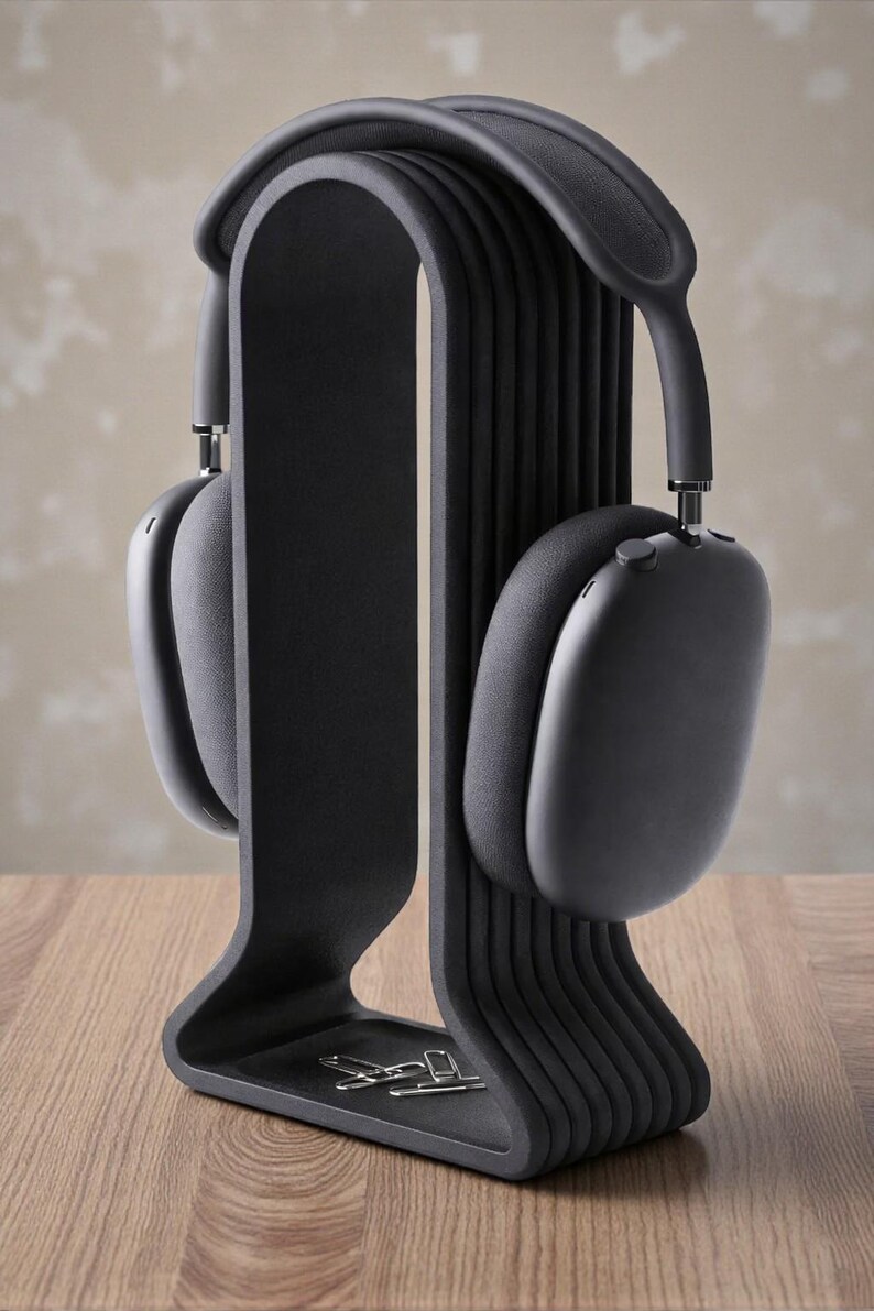 May include: Black over-ear headphones resting on a black headphone stand. The stand has a curved design with a small tray at the base. The headphones and stand are on a wooden surface.