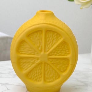 May include: A bright yellow ceramic vase shaped like a lemon slice. The vase has a round body with a narrow neck and a flat base. The lemon slice design is embossed on the front and back of the vase, with the details of the lemon segments clearly visible.