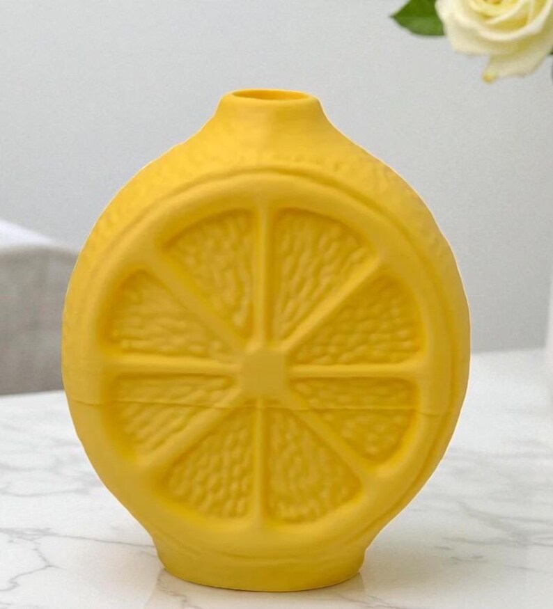 May include: A bright yellow ceramic vase shaped like a lemon slice. The vase has a round body with a narrow neck and a flat base. The lemon slice design is embossed on the front and back of the vase, with the details of the lemon segments clearly visible.