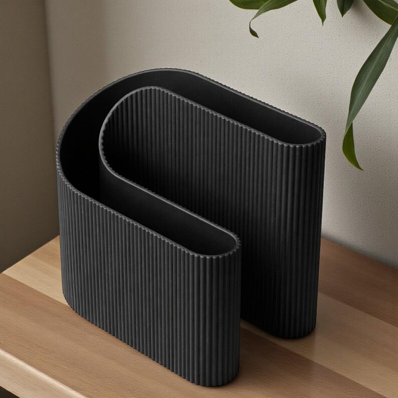 May include: Two black, modern vases with a ribbed texture. The vases have a unique, curved design, and are displayed on a wooden surface. The vases are empty and ready to be filled with flowers or other decorative items.