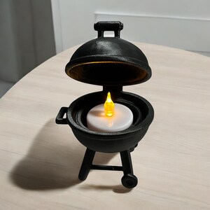 May include: A black miniature barbecue grill with a lid, handle, and a small wheel. Inside the grill is a white candle with a yellow flame. The grill is sitting on a light-colored surface.