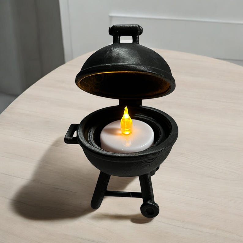 May include: A black miniature barbecue grill with a lid, handle, and a small wheel. Inside the grill is a white candle with a yellow flame. The grill is sitting on a light-colored surface.