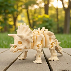 Może przedstawiać: Kremowy, drukowany w 3D model szkieletu Triceratopsa. Dinozaur ma trzy rogi, kołnierz i szczeg&oacute;łową strukturę kości. Model jest prezentowany na drewnianej powierzchni z rozmytym zielonym tłem.