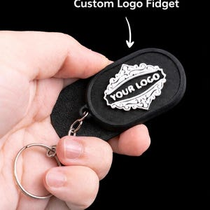 May include: Black custom logo fidget keychain. The keychain features a black oval-shaped fidget with a white logo design and the text "YOUR LOGO". The text "Custom Logo Fidget" is above the fidget.
