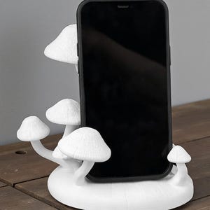 May include: A white, mushroom-shaped phone stand holding a black smartphone. The stand features multiple mushroom caps and stems, creating a whimsical and functional design. The stand is made of a solid white material and sits on a wooden surface.