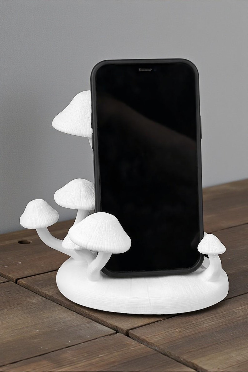 May include: A white, mushroom-shaped phone stand holding a black smartphone. The stand features multiple mushroom caps and stems, creating a whimsical and functional design. The stand is made of a solid white material and sits on a wooden surface.