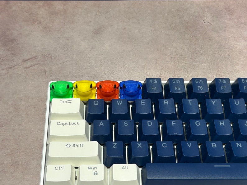 May include: A keyboard with blue keys and white keycaps. Four frog-shaped keycaps are placed on the top row of the keyboard. The frog keycaps are green, yellow, orange, and blue.