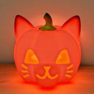 May include: An illuminated orange Halloween decoration in the shape of a pumpkin with cat ears and a carved face. The eyes, nose, and mouth are cut out, allowing light to shine through. It sits on a wooden surface.