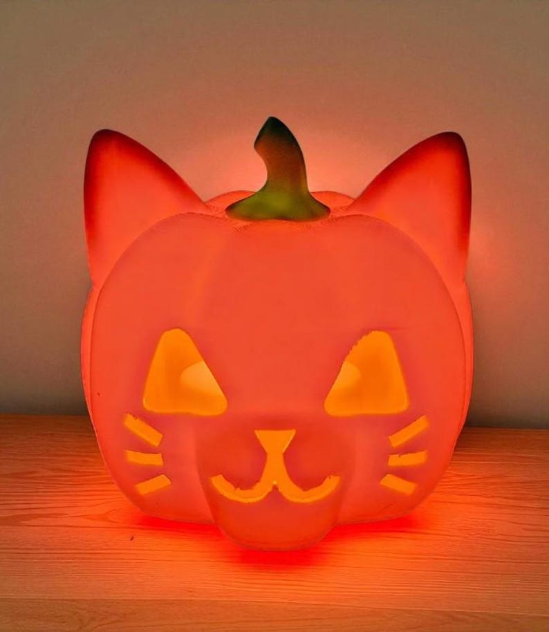May include: An illuminated orange Halloween decoration in the shape of a pumpkin with cat ears and a carved face. The eyes, nose, and mouth are cut out, allowing light to shine through. It sits on a wooden surface.