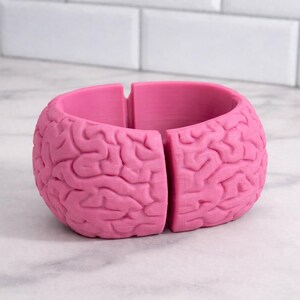 May include: A vibrant pink bracelet, designed to resemble a brain. The bracelet is split into two sections, showcasing the detailed surface of the brain. The item is made of a solid material and rests on a white surface.