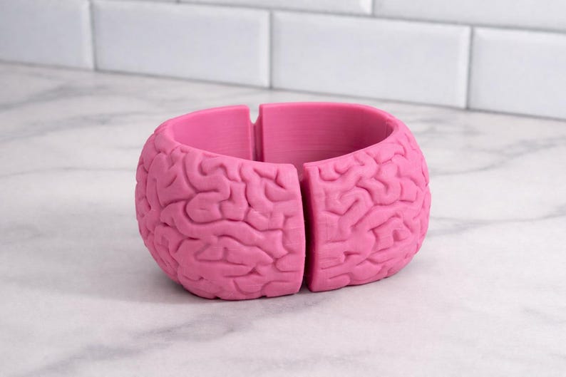 May include: A vibrant pink bracelet, designed to resemble a brain. The bracelet is split into two sections, showcasing the detailed surface of the brain. The item is made of a solid material and rests on a white surface.