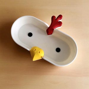 Op de afbeelding: Een witte keramische serveerschaal met een kippenontwerp. De schaal heeft een rode kam, een gele snavel en twee zwarte ogen. De ovale schaal staat op een houten oppervlak.