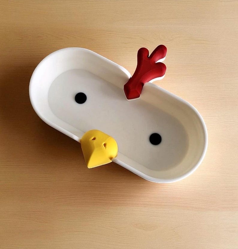 Op de afbeelding: Een witte keramische serveerschaal met een kippenontwerp. De schaal heeft een rode kam, een gele snavel en twee zwarte ogen. De ovale schaal staat op een houten oppervlak.