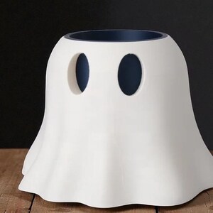 May include: A white ceramic ghost-shaped container with two oval cutouts for eyes. The interior rim is dark blue. The container has a rounded body and a flared, ruffled base, suggesting a Halloween or spooky theme.
