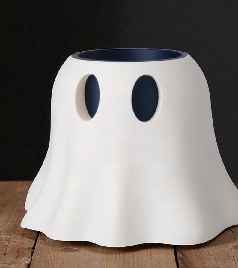 May include: A white ceramic ghost-shaped container with two oval cutouts for eyes. The interior rim is dark blue. The container has a rounded body and a flared, ruffled base, suggesting a Halloween or spooky theme.