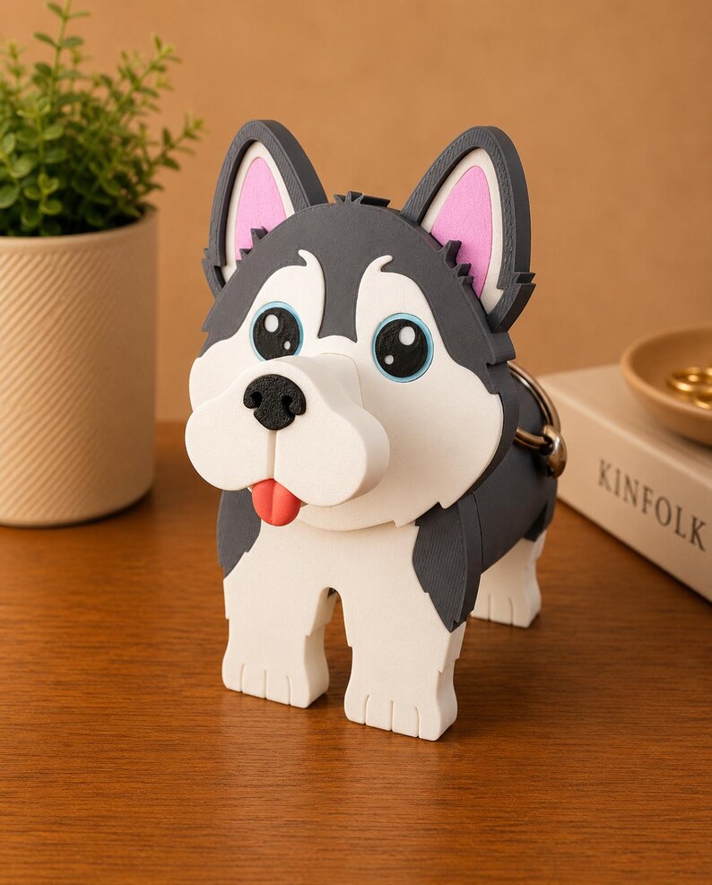 May include: A whimsical, cartoon-style Husky dog figurine. The dog has a gray and white coat, large blue eyes, and a red tongue. The figurine is standing on a wooden surface. A small plant in a beige pot is in the background.