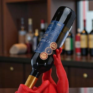 May include: A red frog-shaped wine bottle holder supports a dark wine bottle. The bottle has a blue label with gold accents and several round stickers. The holder is on a wooden surface, with a blurred bar background.