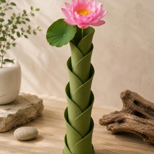May include: A green, spiral-shaped vase holds a pink lotus flower with a yellow center and a green leaf. The vase has a textured surface and is made of a material that resembles folded leaves. The flower is in full bloom, adding a pop of color.