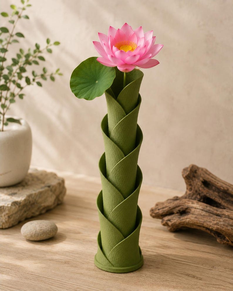 May include: A green, spiral-shaped vase holds a pink lotus flower with a yellow center and a green leaf. The vase has a textured surface and is made of a material that resembles folded leaves. The flower is in full bloom, adding a pop of color.