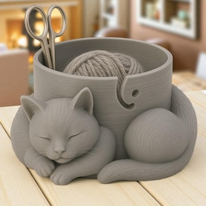 May include: A gray cat-shaped yarn bowl with a ball of yarn inside. The bowl has a slot for the yarn and holds a pair of silver scissors. The cat is curled up, appearing to be sleeping. The bowl is on a light wood surface.
