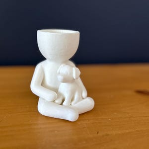 Puede incluir: Figura de cer&aacute;mica blanca que representa a una persona sentada con un perro peque&ntilde;o. La figura tiene un dise&ntilde;o minimalista con una cabeza hueca, y el perro est&aacute; en el regazo de la persona. Sobre una superficie de madera.