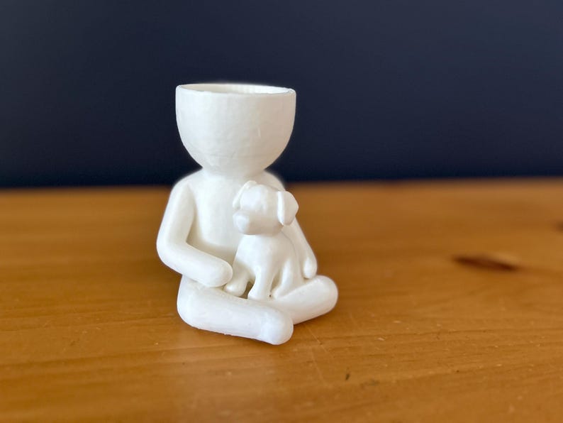 Puede incluir: Figura de cer&aacute;mica blanca que representa a una persona sentada con un perro peque&ntilde;o. La figura tiene un dise&ntilde;o minimalista con una cabeza hueca, y el perro est&aacute; en el regazo de la persona. Sobre una superficie de madera.