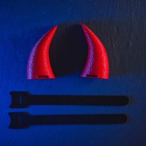 May include: Two red devil horns on a blue background. The horns are made of a soft, flexible material and have a pointed tip. The horns cast a shadow on the surface below.