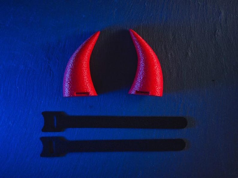 May include: Two red devil horns on a blue background. The horns are made of a soft, flexible material and have a pointed tip. The horns cast a shadow on the surface below.