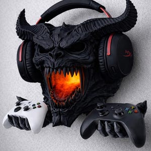 Può includere: Un supporto nero per cuffie e controller da gioco a tema demone. Il supporto presenta un volto dettagliato e minaccioso con corna, una bocca luminosa e mani tese che tengono un controller bianco e uno nero. Le cuffie sono nere con dettagli rossi.