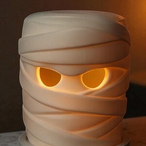 K&ouml;nnte beinhalten: Cremefarbene mumienf&ouml;rmige Lampe mit leuchtend orangefarbenen Augen. Die Lampe ist mit strukturierten B&auml;ndern umwickelt, was ihr ein realistisches Aussehen verleiht. Sie steht auf einem runden, hellgrauen Sockel und ist eine gruselige Halloween-Dekoration.
