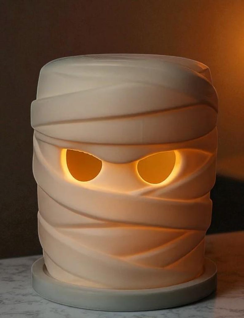 K&ouml;nnte beinhalten: Cremefarbene mumienf&ouml;rmige Lampe mit leuchtend orangefarbenen Augen. Die Lampe ist mit strukturierten B&auml;ndern umwickelt, was ihr ein realistisches Aussehen verleiht. Sie steht auf einem runden, hellgrauen Sockel und ist eine gruselige Halloween-Dekoration.