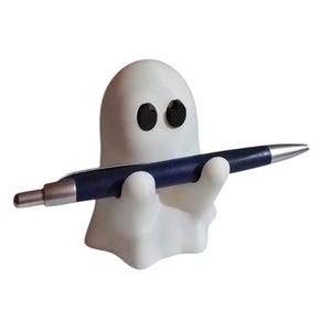 May include: A white ghost-shaped pen holder with black eyes, holding a blue and silver pen. The pen holder is a whimsical desk accessory, perfect for adding a touch of fun to any workspace. The pen is approximately 5.5 inches long.