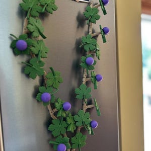 May include: A decorative vine-shaped magnet with green leaves and purple grape-like spheres. The vine is attached to a refrigerator door. The leaves and grapes are made of a plastic-like material.