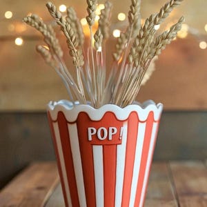 May include: A ceramic popcorn bucket-shaped vase with red and white stripes, holding dried wheat stalks. The word "POP!" is displayed on the front. The vase has a scalloped white rim and sits on a wooden surface.