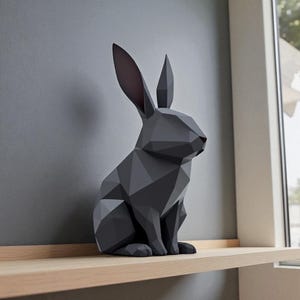 May include: A geometric, dark gray rabbit sculpture with a red nose and pink inner ears. The modern art piece is displayed on a light wood shelf against a gray wall. The rabbit is a decorative item.