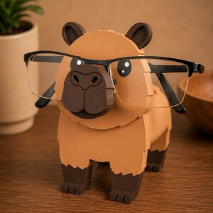 May include: A whimsical, tan capybara-shaped glasses holder with dark brown accents. The holder features a detailed nose and ears, and is designed to hold a pair of black-framed glasses. The glasses are positioned as if the capybara is wearing them.