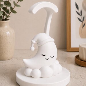 May include: A white night light shaped like a crescent moon wearing a sleeping cap, resting on a cloud-like base. The lamp has a curved arm and a round base. The moon has closed eyes. The lamp is designed for a nursery or child's room.