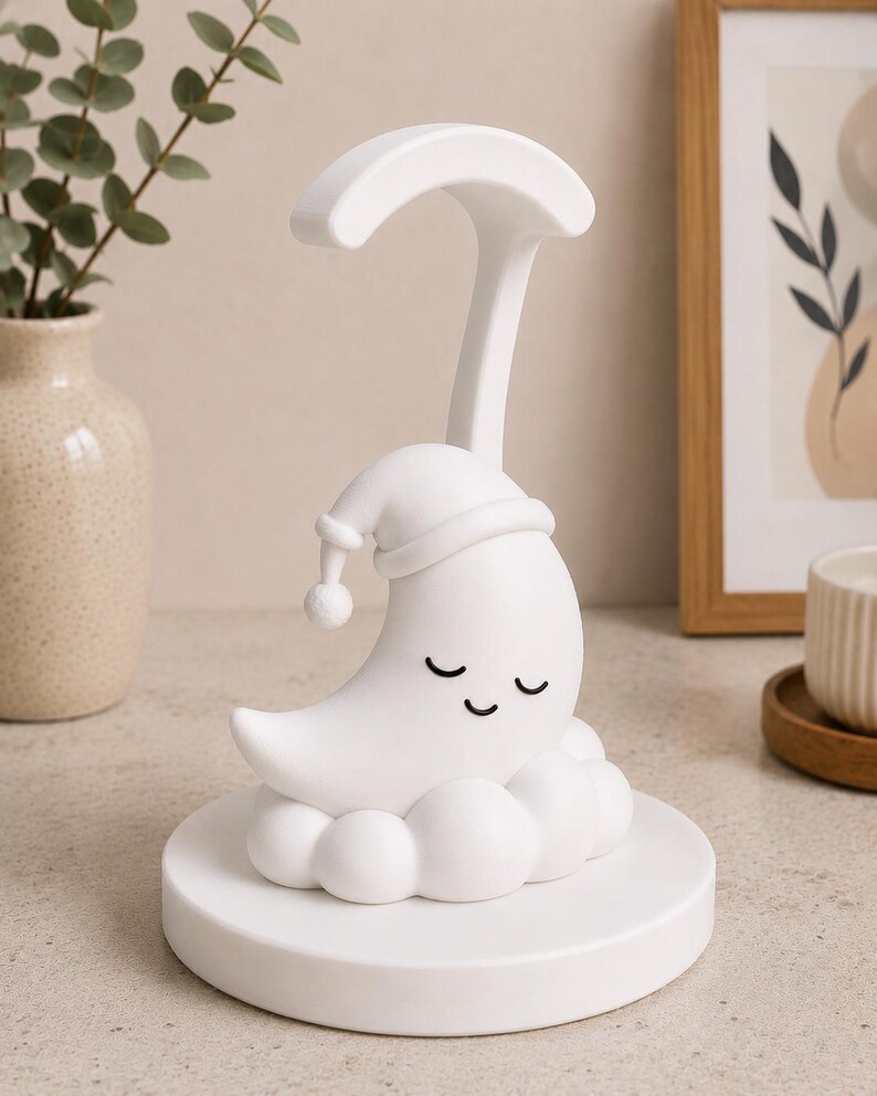 May include: A white night light shaped like a crescent moon wearing a sleeping cap, resting on a cloud-like base. The lamp has a curved arm and a round base. The moon has closed eyes. The lamp is designed for a nursery or child's room.